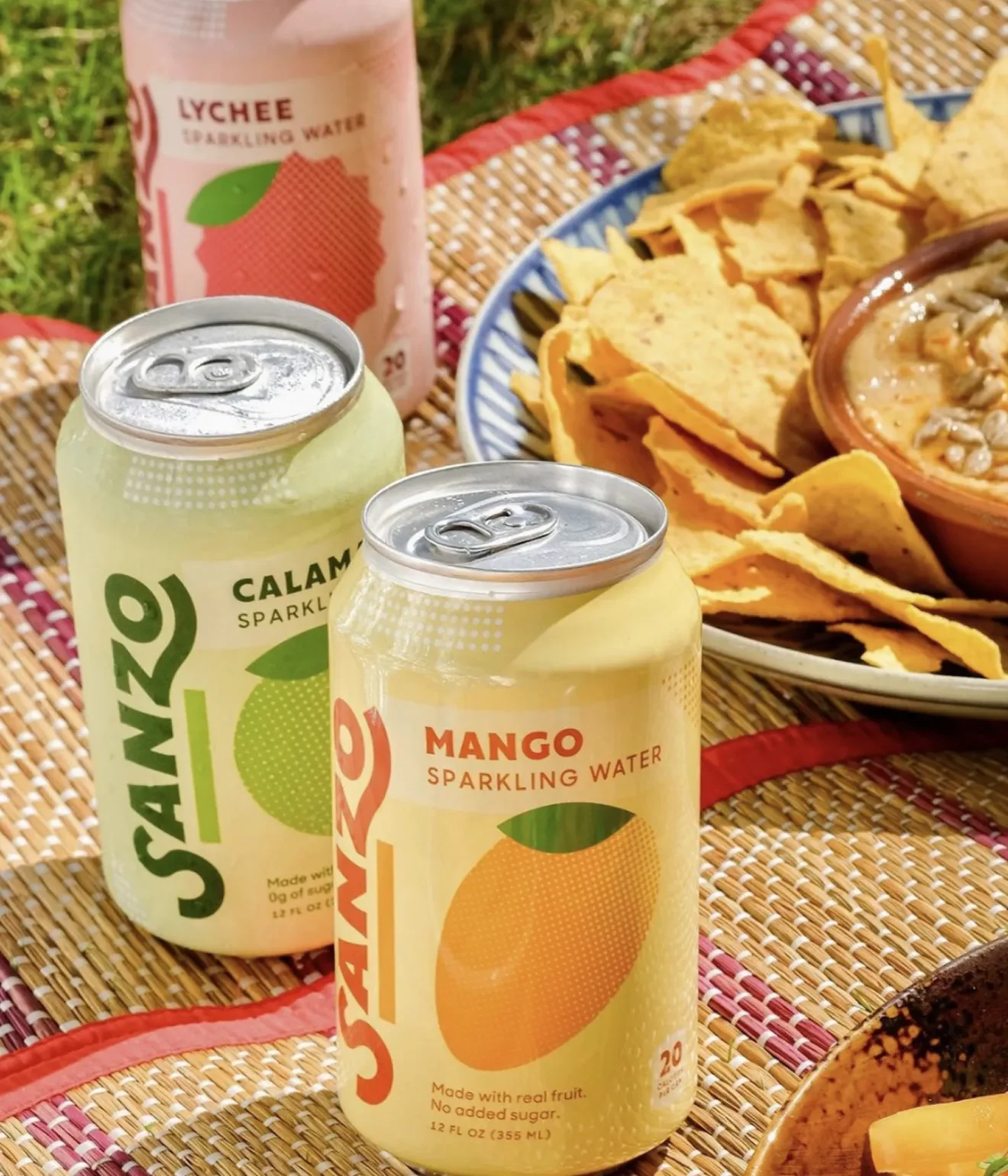 Sanzo sparkling water cans in mango and calamansi on a picnic table with chips and salsa.